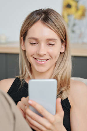 Cheerful girl with long blond hair looking at her friend on smartphone screen
