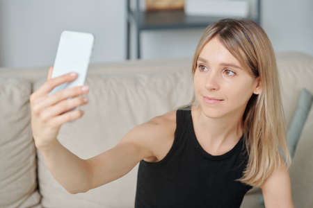 Young smiling blond female in black vest looking in smartphone camera