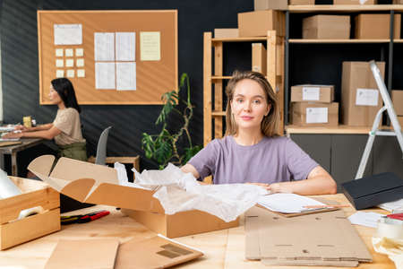 Young female worker of online shop office sitting by table in front of camera