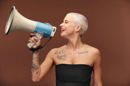 Happy young blond female with tattoos on arm, chest and neck holding megaphone