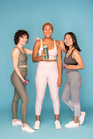 Three young laughing sportswomen holding bottle of water and dumbbellsの写真素材
