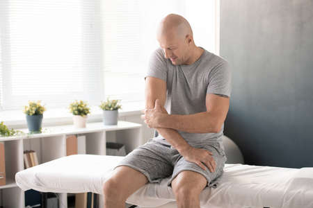 Bald man touching painful elbow and trying to massage it before exercising