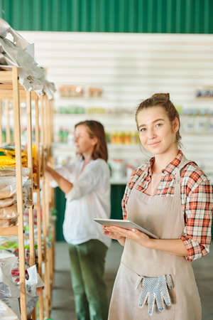 Young pretty female assistant of gardening supermarket in workwear using tablet