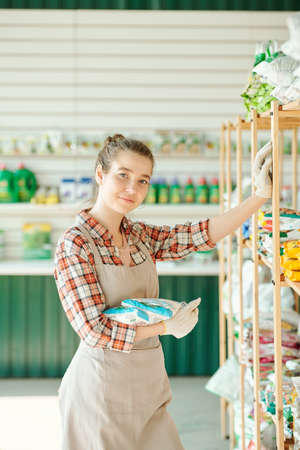 Young pretty assistant of gardening shop with packs of granulated fertilizer