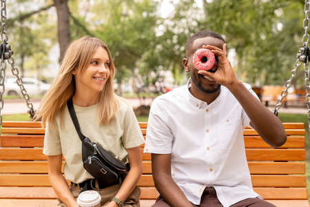 Happy young blond woman with drink looking at funny and joyful African man