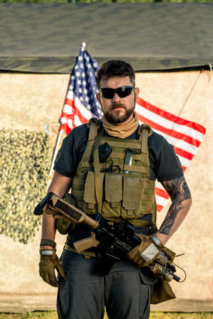 Portrait of serious brutal bearded man in vest with radio device holding riffle against American...