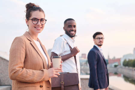 Portrait of positive adult students standing in row, focus on attractive girl in glasses drinking...