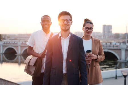 Team of business people standing together against city bridge in sunlight, young leader in glasses...
