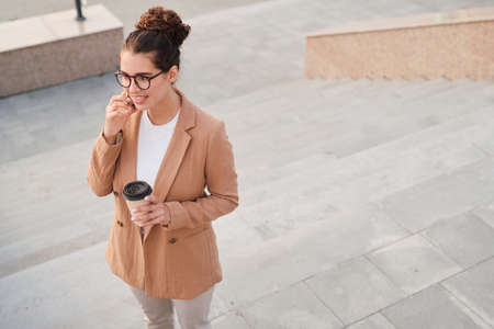Above view of account manager in jacket drinking coffee and talking to client by phone being on moveの写真素材
