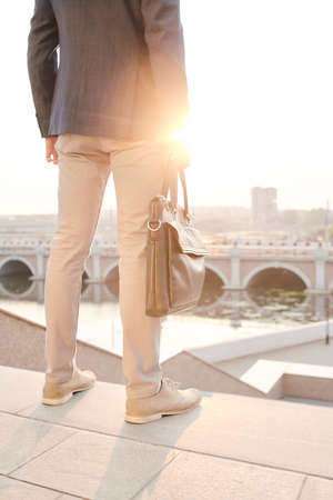 Rear view of unrecognizable businessman with briefcase contemplating city while leaving office, sunlight effectの写真素材