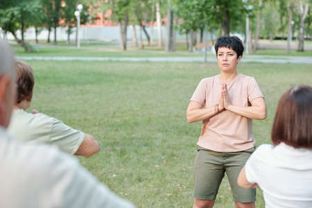Focused yoga teacher making Namaste gesture while meditating with students in summer parkの写真素材