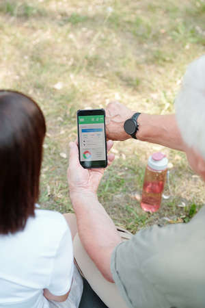 Over shoulder view of senior man synchronizing fitness tracker with smartphone while resting with girlfriend after trainingの写真素材