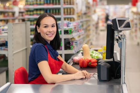 Cheerful woman working in supermarket sitting at cash desk looking at camera smilingの写真素材