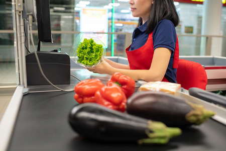 Asian woman wearing red apron sitting at cash desk beeping vegetablesの写真素材
