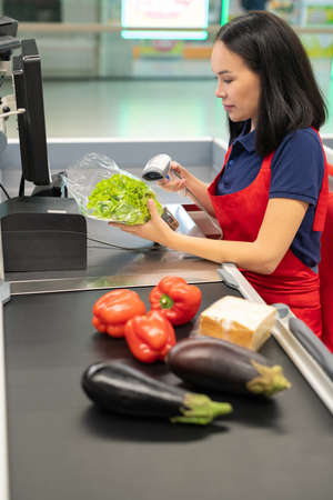 Vertical shot of attractive Asian cashier wearing red apron selling fresh vegetables to customer