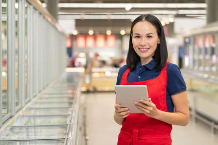 Attractive Asian woman wearing uniform with red apron working in supermarket holding digital tablet...