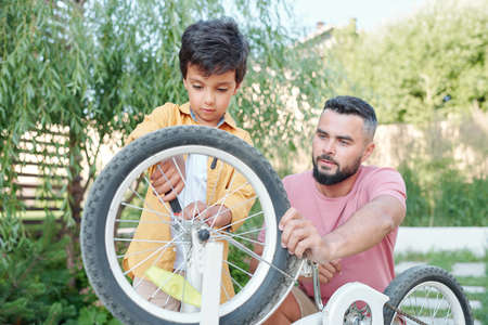 Young father holding bicycle while his little son trying to fix its wheel, horizontal medium shotの写真素材