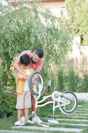 Loving father and his son spending time together outdoors repairing bicycle on sunny day, vertical...