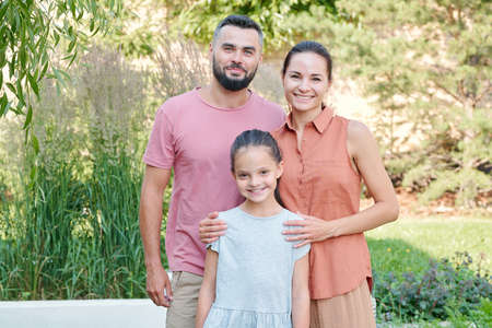 Family portrait of father, mother and daughter in casual summer outfits standing together outdoors in park looking at camera smilingの写真素材