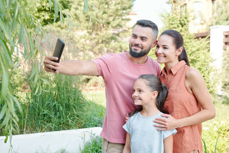 Young adult man holding smartphone taking selfie shot of him and his wife and daughter in backyard on sunny summer dayの写真素材