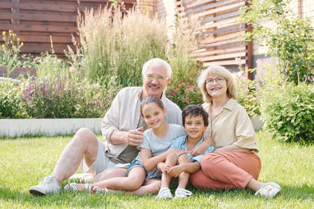 Group portrait of cheerful grandma, grandpa and two grandchildren sitting together on lawn in backyard looking at camera smilingの写真素材