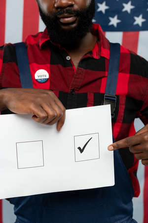 Young African-american repairman with vote insignia pointing at ballot paper