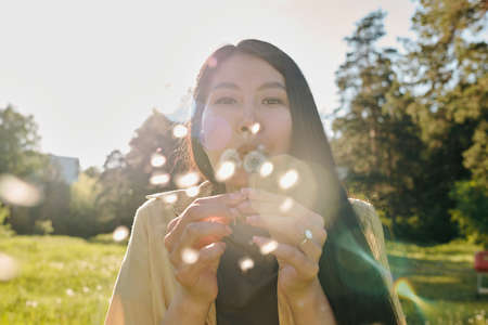 Young Asian woman with long dark hair blowing at dandelion while having funの写真素材