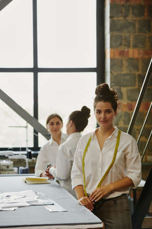 Young serious seamstress with measuring tape on her neck standing by tableの写真素材