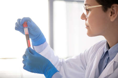 Gloved female scientist in whitecoat looking at flask with red liquid substance