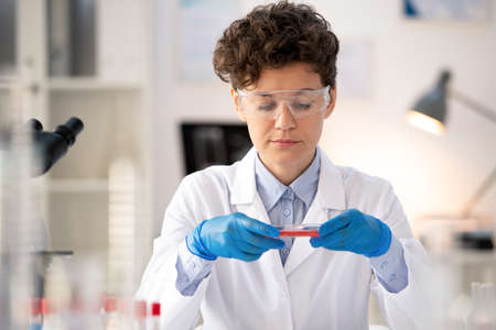 Contemporary female scientist in whitecoat and gloves sitting by workplace