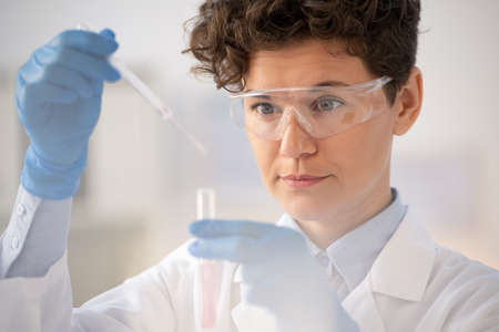 Gloved laboratory worker in eyeglasses dropping fluid from pipette into flaskの写真素材