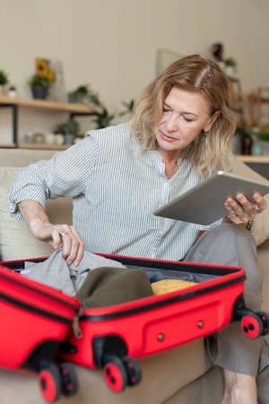 Mature blond woman with touchpad putting clothes and other stuff into suitcaseの写真素材