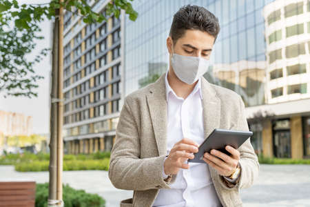 Young attractive elegant businessman with protective mask using touchpadの写真素材