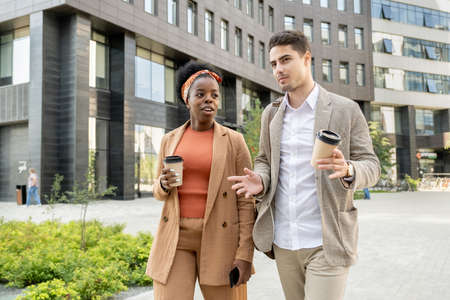 Young African woman having drink while moving along park next to male colleagueの写真素材