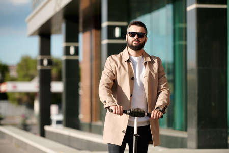 Serious bearded businessman in beige trenchcoat and sunglasses riding scooter