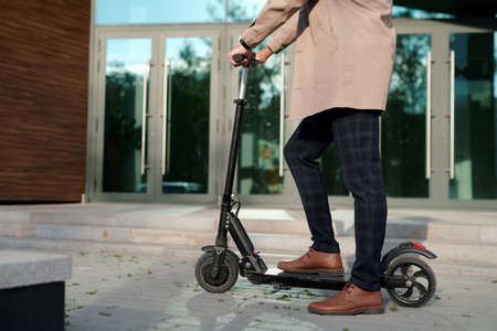 Low section of businessman in trenchcoat and pants holding by handles of scooter