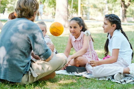Curious multi-ethnic schoolgirls holding planet models on sticks while discussing astronomical bodies with teacher in parkの写真素材