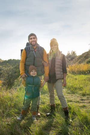 Young cheerful parents and their little son with backpacks standing on grass