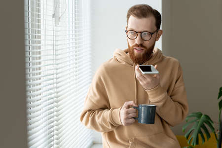 Young contemporary man in casualwear and eyeglasses recording voice message