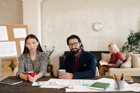 Two young elegant colleagues with mugs of coffee or tea sitting by workplaceの写真素材