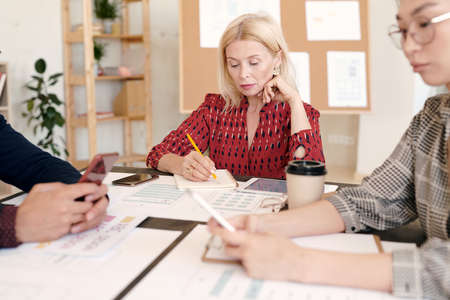 Mature blond female mobile application developer making notes in notebook