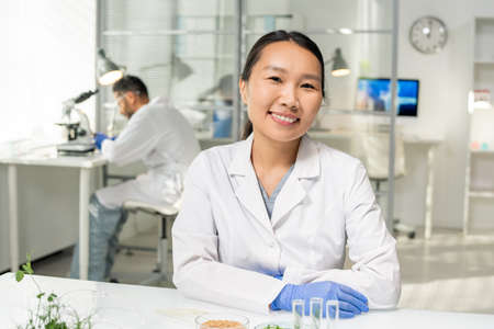 Young successful Asian female researcher in whitecoat and gloves looking at youの写真素材