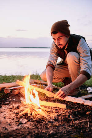 Serious young bearded hiker in cap putting wood while setting fire near coastline at sunsetの写真素材