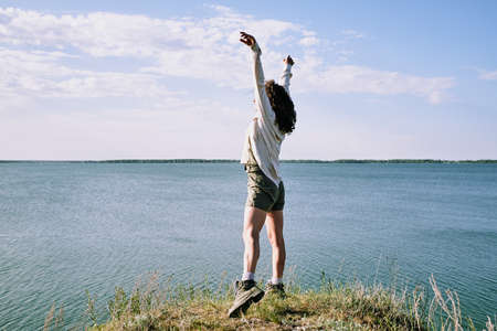 Carefree young female hiker with curly hair standing with raised arms and catching sunlight at...
