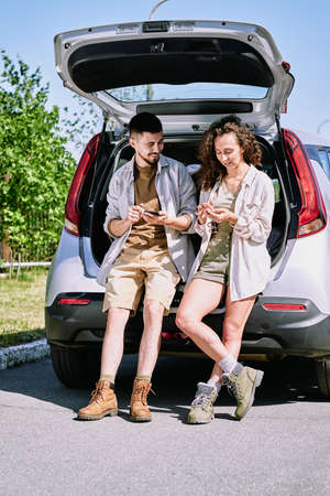 Positive active young hikers in boots leaning on car trunk and discussing hiking route using phone appsの写真素材
