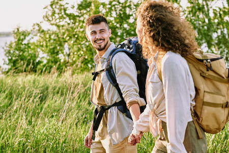 Positive young couple with backpacks holding hands and looking at each other while enjoying hike in...