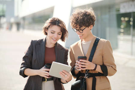 Two happy young colleagues with drinks looking at screen of digital tabletの写真素材