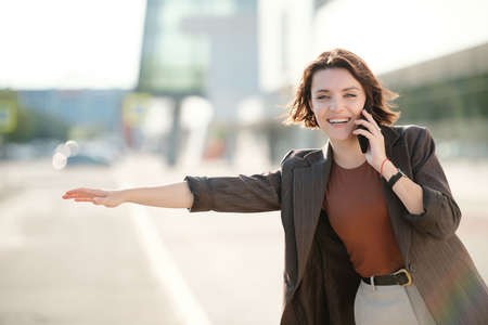 Young cheerful brunette businesswoman with smartphone by ear catching taxiの写真素材