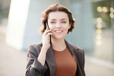 Young smiling brunette businesswoman with smartphone by ear consulting client on the phone against contemporary architecture exteriorの写真素材