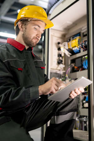 Young bearded engineer in workwear and hardhat checking settings of machine